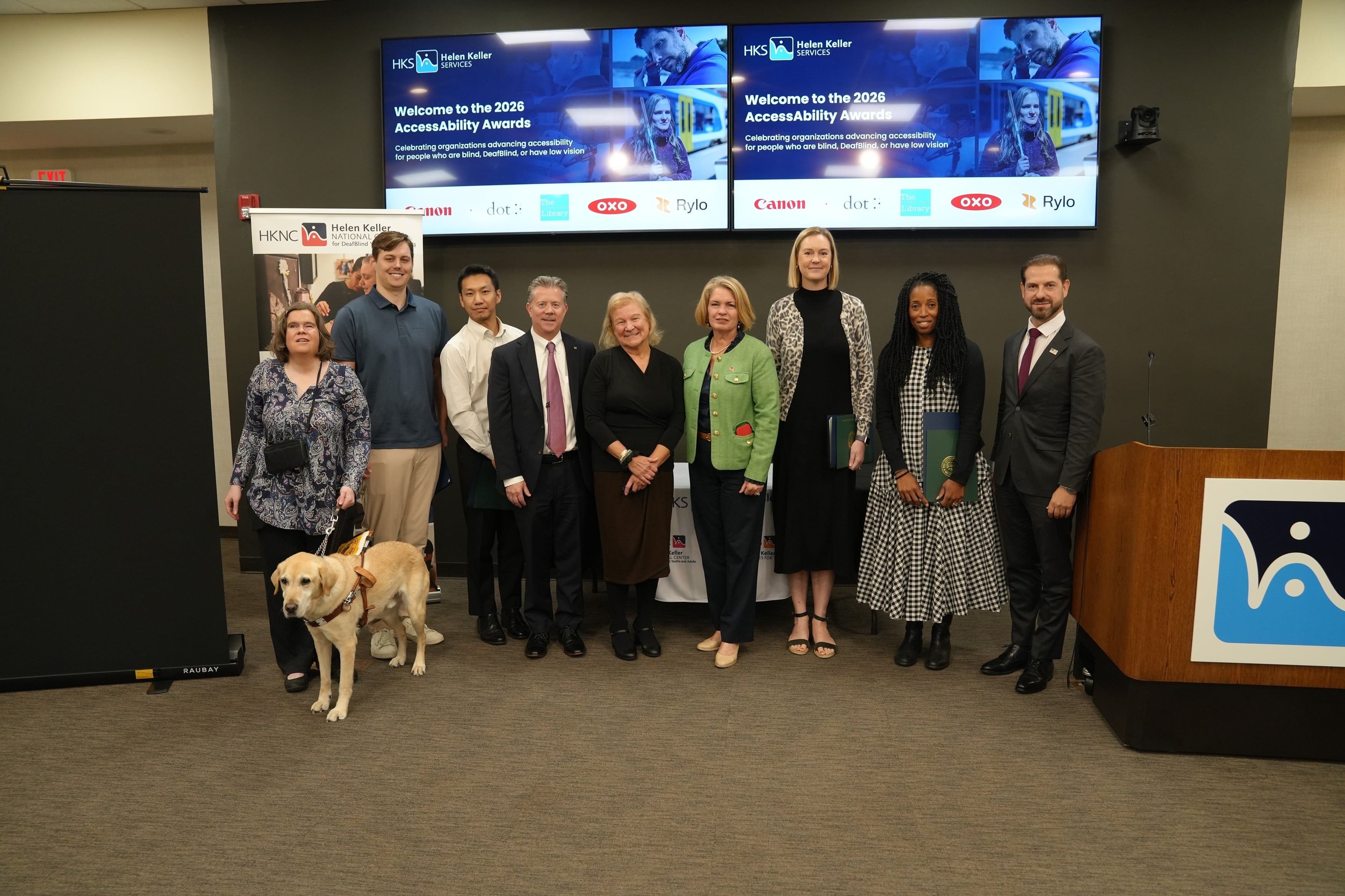 A group photo at the 2026 AccessAbility Awards at the Helen Keller National Center. Two screens behind the group read “Welcome to the 2026 AccessAbility Awards: Celebrating organizations advancing accessibility for people who are blind, DeafBlind, or have low vision,” above the logos of Canon, Dot, The Indianapolis Public Library, OXO, and Rylo. From left: Helen Keller Services Accessibility Specialist Megan Dausch with her yellow Labrador guide dog; three representatives of the 2026 AccessAbility Award honorees; Sue Ruzenski, CEO of Helen Keller Services; Town of North Hempstead Supervisor Jennifer DeSena; two more honoree representatives; and New York State Assemblyman Daniel Norber