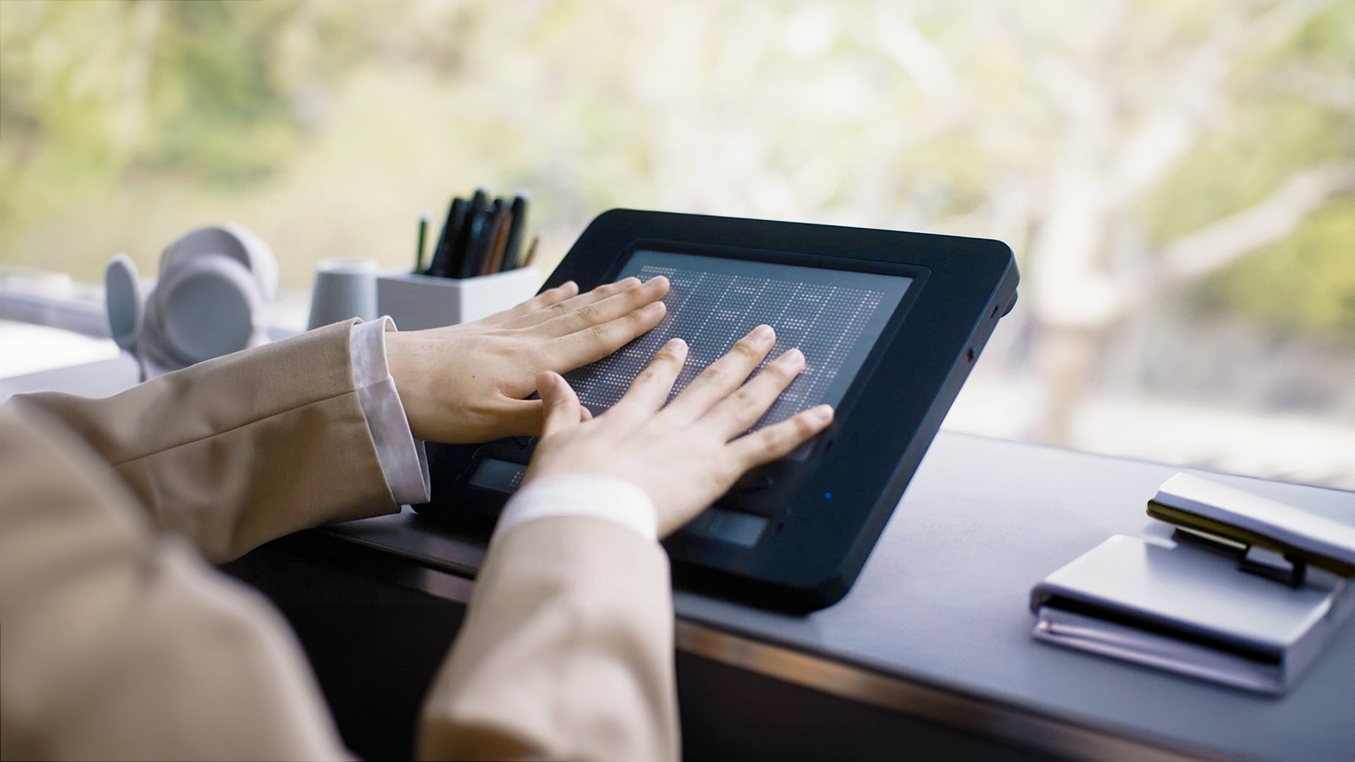 The Dot Pad X is placed on a table. Two hands are on the screen reading a tactile graphic
